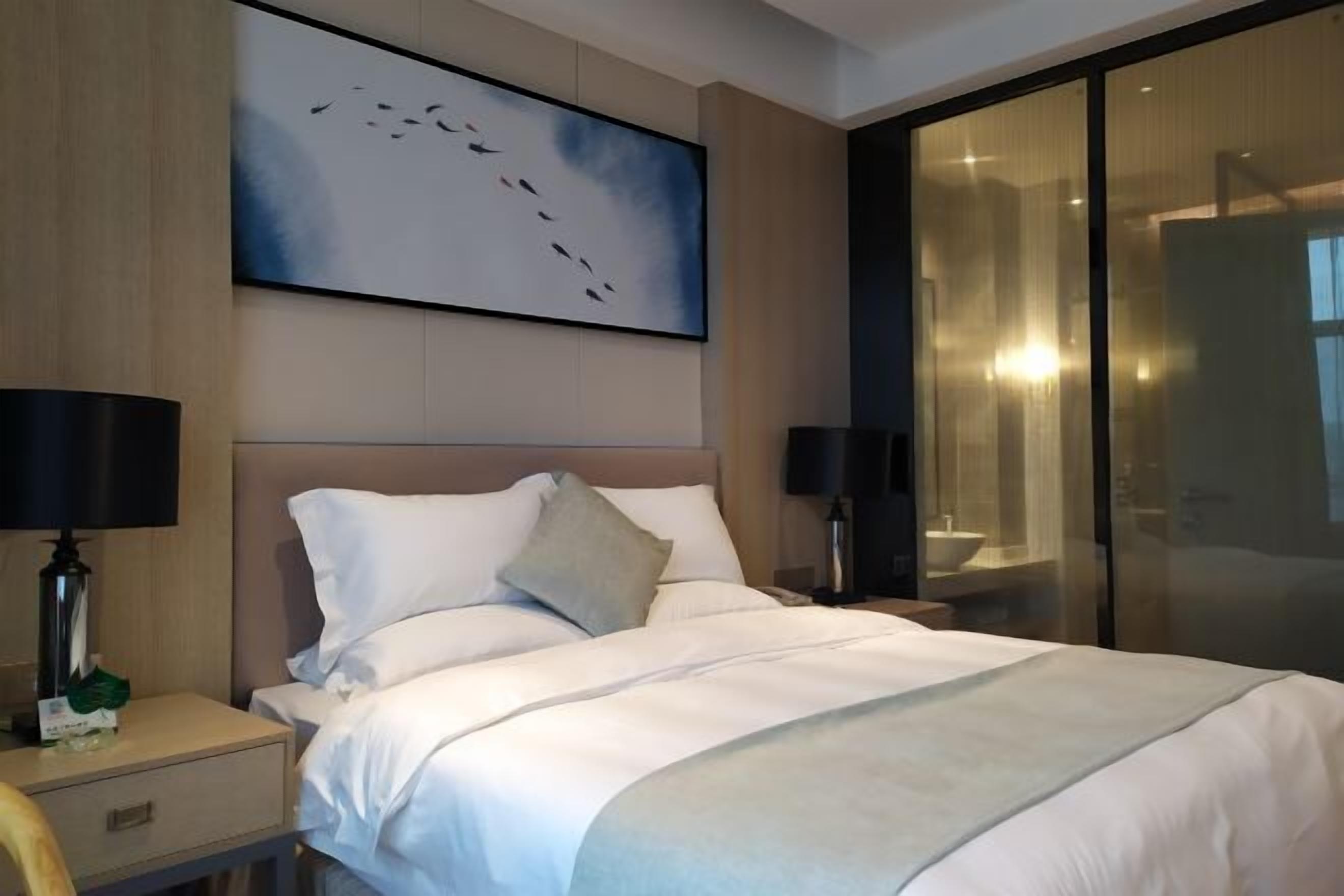Habitación de hotel con una cama grande, dos mesitas de noche, una mesilla auxiliar, un televisor montado en la pared y una puerta de vidrio que da al baño.