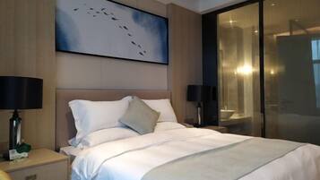 Habitación de hotel con una cama grande, dos mesitas de noche, una mesilla auxiliar, un televisor montado en la pared y una puerta de vidrio que da al baño.