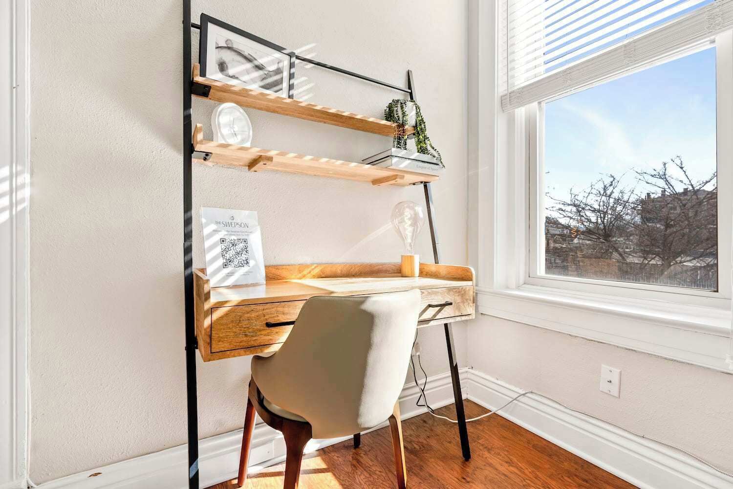 Un bureau d’angle avec une chaise, une étagère murale avec une horloge et un tableau encadré, et une fenêtre donnant sur des arbres et des immeubles.