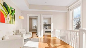 Un salón amplio con un sofá blanco, una lámpara de pie y un cuadro abstracto de gran tamaño. Una escalera de madera conduce a un balcón con barandilla.