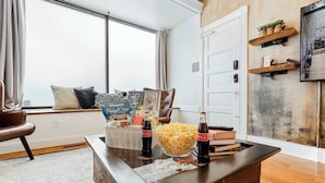 Una sala de estar con una mesa de centro que sostiene aperitivos, bebidas y una revista. Hay una ventana con cortinas, una estantería y una puerta con un perchero.