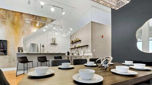 Un comedor moderno con una larga mesa de madera dispuesta para una comida, una cocina al fondo y un amplio ventanal que proporciona luz natural.