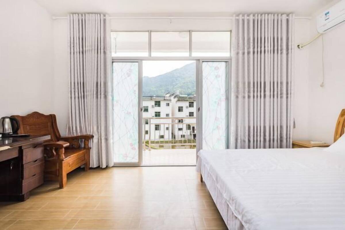 A hotel room with a bed, a wooden desk, a chair, and a view of buildings through a glass door.