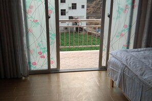 Un dormitorio con cama, puerta corrediza de vidrio que da a un balcón y vista a un edificio y vegetación.