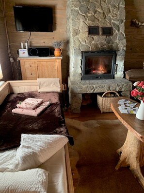 Um quarto acolhedor com lareira de pedra, televisão de tela plana na parede, armário de madeira, cama com cobertura marrom escura e uma pequena mesa com um vaso de flores.
