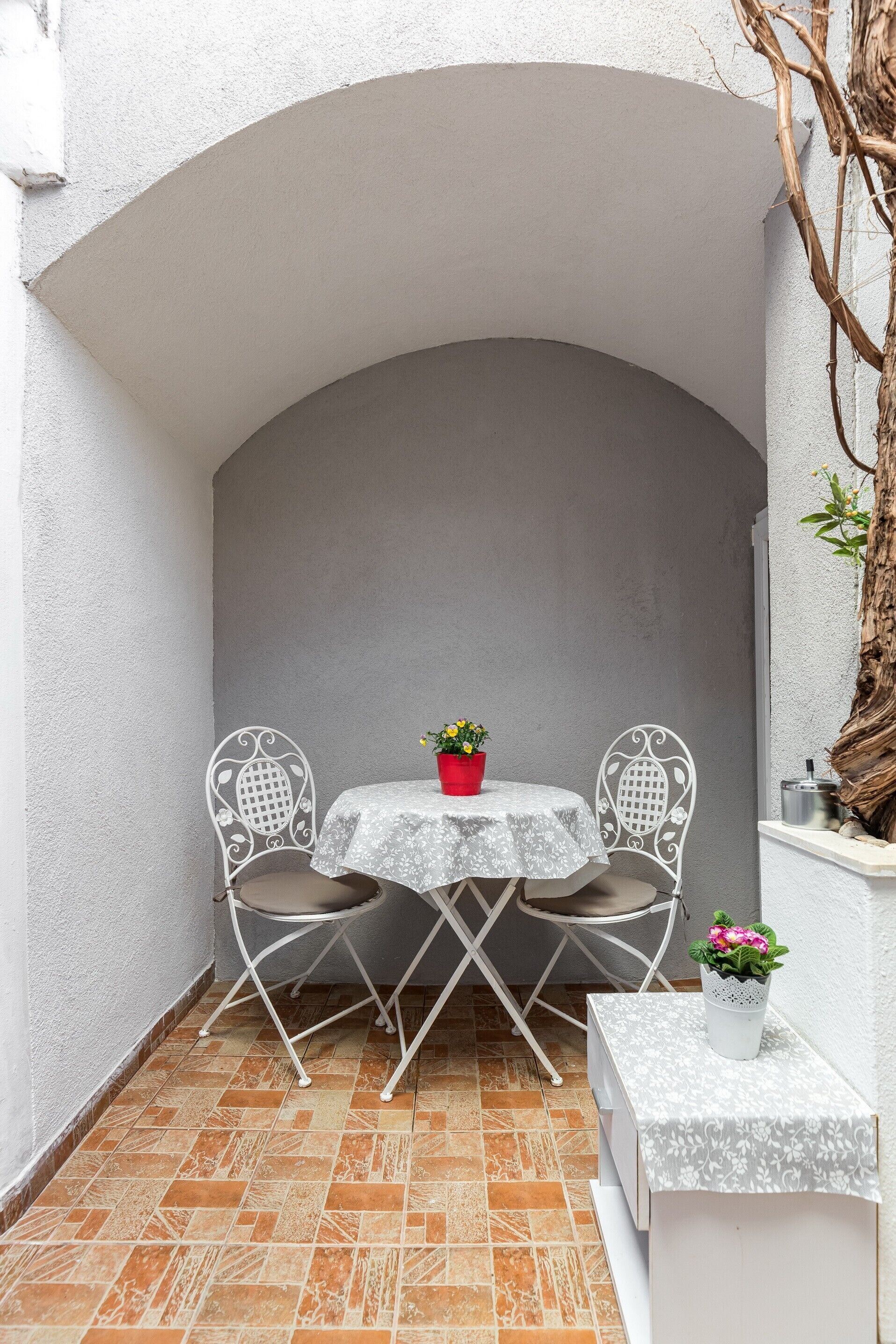 Un pequeño patio al aire libre con una mesa blanca y dos sillas, una planta en maceta y un banco blanco.