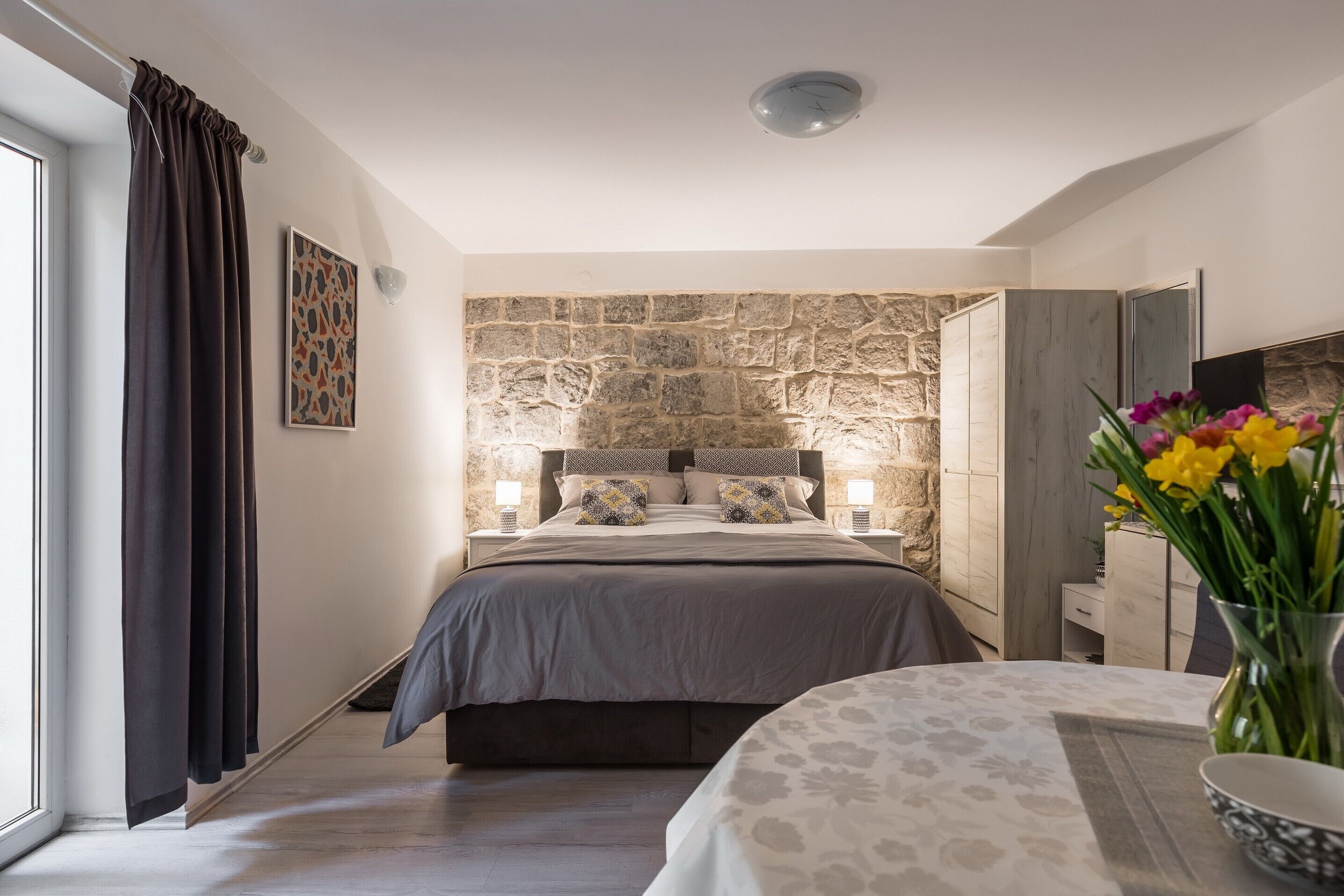 Een slaapkamer met een stenen muur, een bed met een grijze spreidek, twee bedlampen en een vaas met bloemen op een nachtkastje.