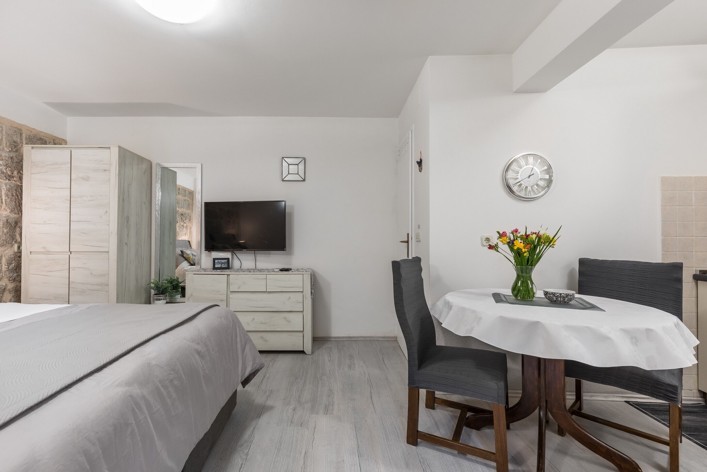 A bedroom with a bed, a dresser, a TV, a dining table with chairs, and a clock on the wall.