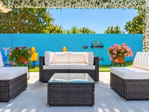 A patio with wicker furniture, a blue wall, and a garden. - Underwater Voyage: Heated Pool, Slide, Arcade, more! (Garden Grove)