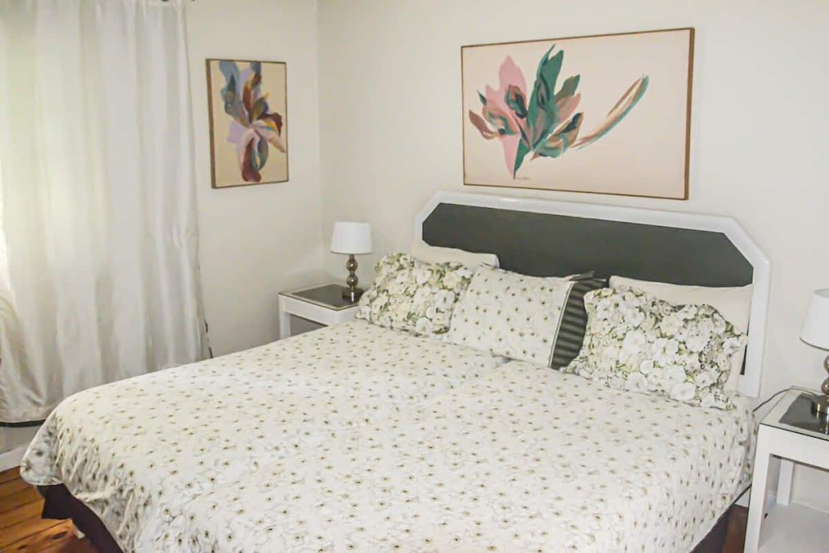 Quarto com cama, cortinas brancas, mesa de cabeceira com abajur e duas obras de arte floral emolduradas na parede.