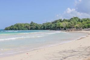 A beach with clear turquoise water, sandy shore, and a forested hill in the background.