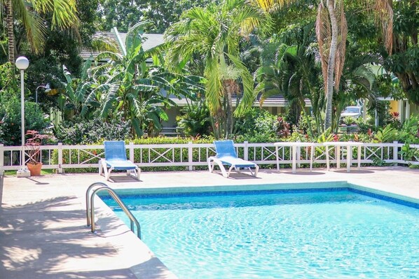 A swimming pool with two blue lounge chairs, surrounded by lush tropical plants and a white fence.