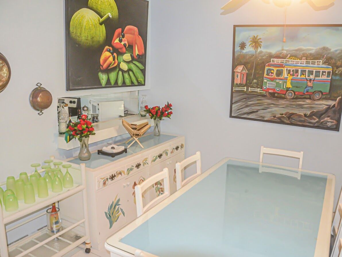 A room with a glass-top table, a white cabinet with drawers, a shelf with green bottles, and two paintings on the wall.