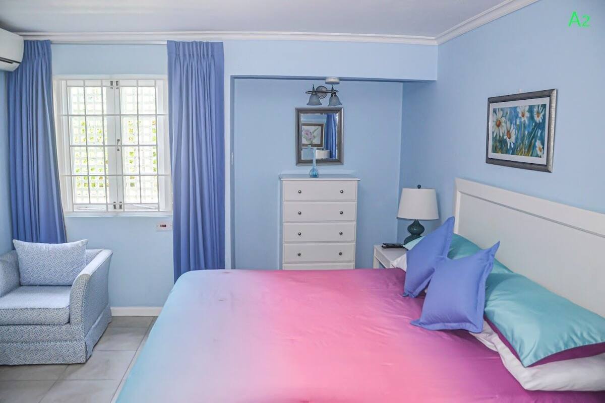 Quarto com cama, mesa de cabeceira, cômoda, cadeira e janela com cortinas.