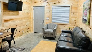Ein gemütliches Blockhaus mit Holzfassade, einer grauen Tür, einem Fenster mit Jalousien, einem schwarzen Ledersofa, einem gemusterten Sessel, einem kleinen Tisch und einem an der Wand montierten Fernseher.