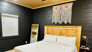 Ein Schlafzimmer mit einem Holz-Kopfteil, einem Bett mit weißer Bettwäsche, einem Wandbehang aus Makramee, einer kleinen Holzablage und einem Nachttisch mit Lampe.
