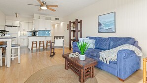 A modern living room with a blue sofa, wooden coffee table, and a ceiling fan. A kitchen with white cabinets and a dining area with stools is visible in the background. - Duckburgh - Ocean view, Updated, 2 min. walk to the beach (Kitty Hawk)