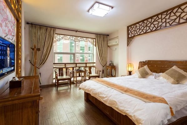 A hotel room with a large bed, a TV on the left, a wooden desk, and a window with curtains.
