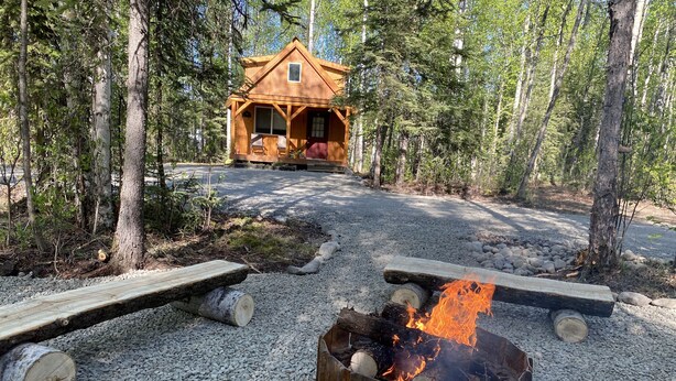 Une cabane rustique avec un foyer au premier plan, entourée de gravier et d’arbres.