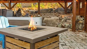 A square fire pit table with a fire burning in the center, surrounded by a wooden bench and a blue chair.