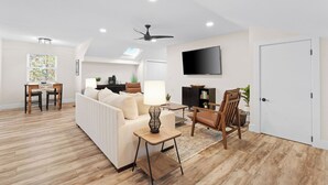 A modern living room with a sofa, coffee table, and chairs, a dining area with a table and chairs, a TV mounted on the wall, and a ceiling fan.