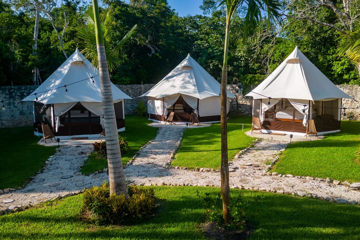Three white tents with attached wooden beds, set on a stone-paved path surrounded by grass and palm trees.