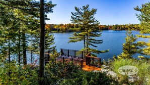 A lakeside cabin with a wooden deck and a view of the lake and surrounding trees. - 6 bed 4 bath cottage on Lake Muskoka, Private, Boathouse, Hot tub! (Gravenhurst)