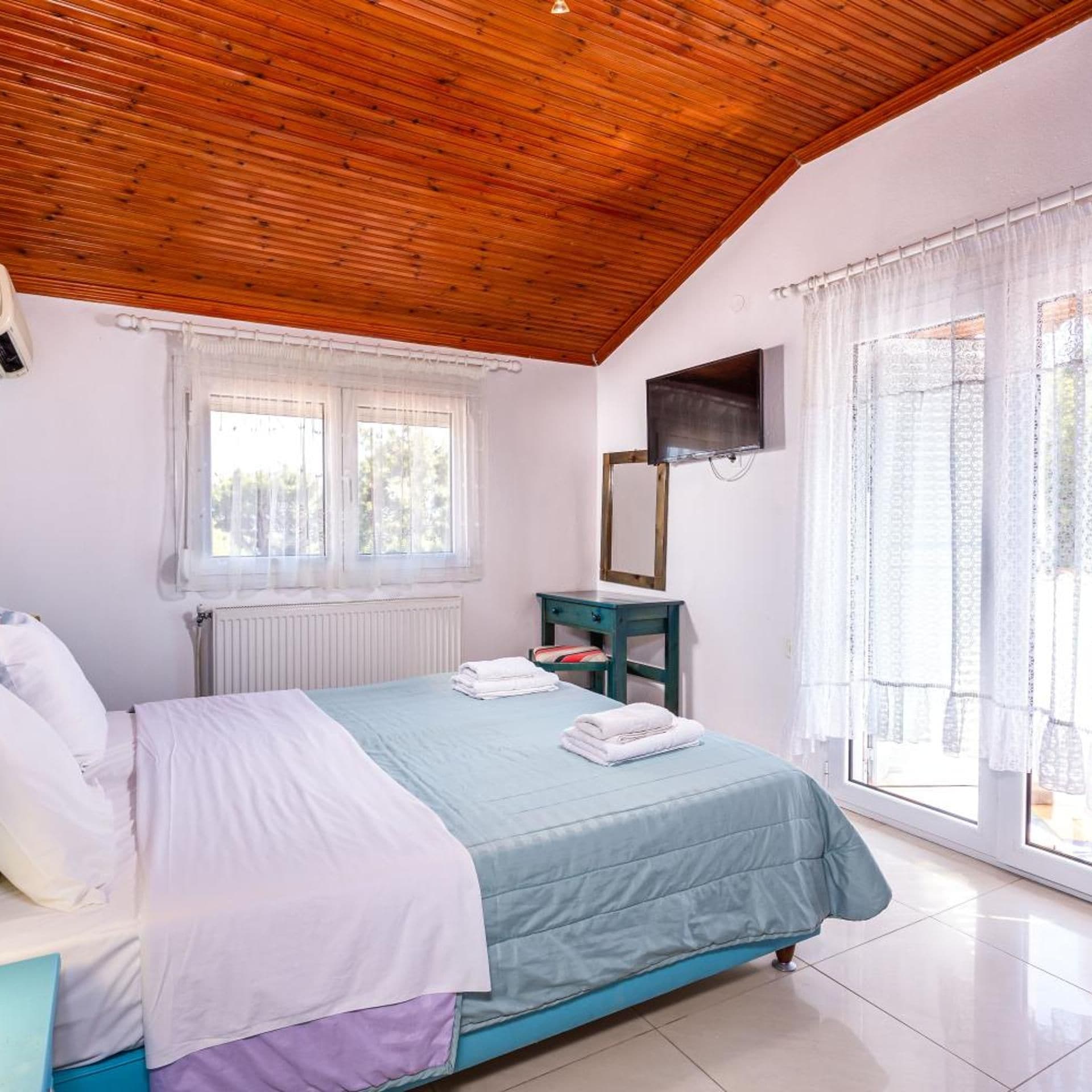 Un dormitorio con techo de madera, una cama con colcha azul y blanca, una mesita de noche con espejo y una ventana con cortinas translúcidas.