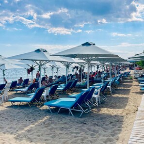 A beach with rows of sun loungers under umbrellas, a wooden walkway, and the sea in the background.