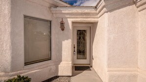 A building with a white stucco exterior, a closed garage door, a glass door with a decorative pattern, and a wall-mounted light fixture. - Beautiful custom home w pool & garage! Perfect snowbird retreat, close to all! (Lake Havasu City)