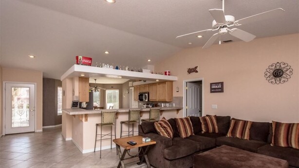 A spacious living room with a brown sectional sofa, a small wooden table, and a ceiling fan. A kitchen area is visible in the background with a bar counter and seating. - Beautiful custom home w pool & garage! Perfect snowbird retreat, close to all! (Lake Havasu City)