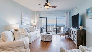 A spacious living room with a large window, a ceiling fan, a flat-screen TV, and a cozy seating area. - The Palms 215B | Spacious, recent updates, beach front! (Orange Beach)