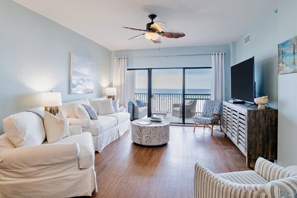 A spacious living room with a large window, a ceiling fan, a flat-screen TV, and a cozy seating area.