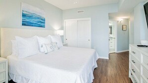 A bedroom with a large bed, white nightstands, a ceiling fan, a door, and a hallway leading to a bathroom. - The Palms 215B | Spacious, recent updates, beach front! (Orange Beach)
