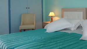 A neatly made bed with a teal bedspread and white pillows, a wooden nightstand with a lamp, and a chair in a room with light blue walls. - Roda Pearl Resort (Corfu)