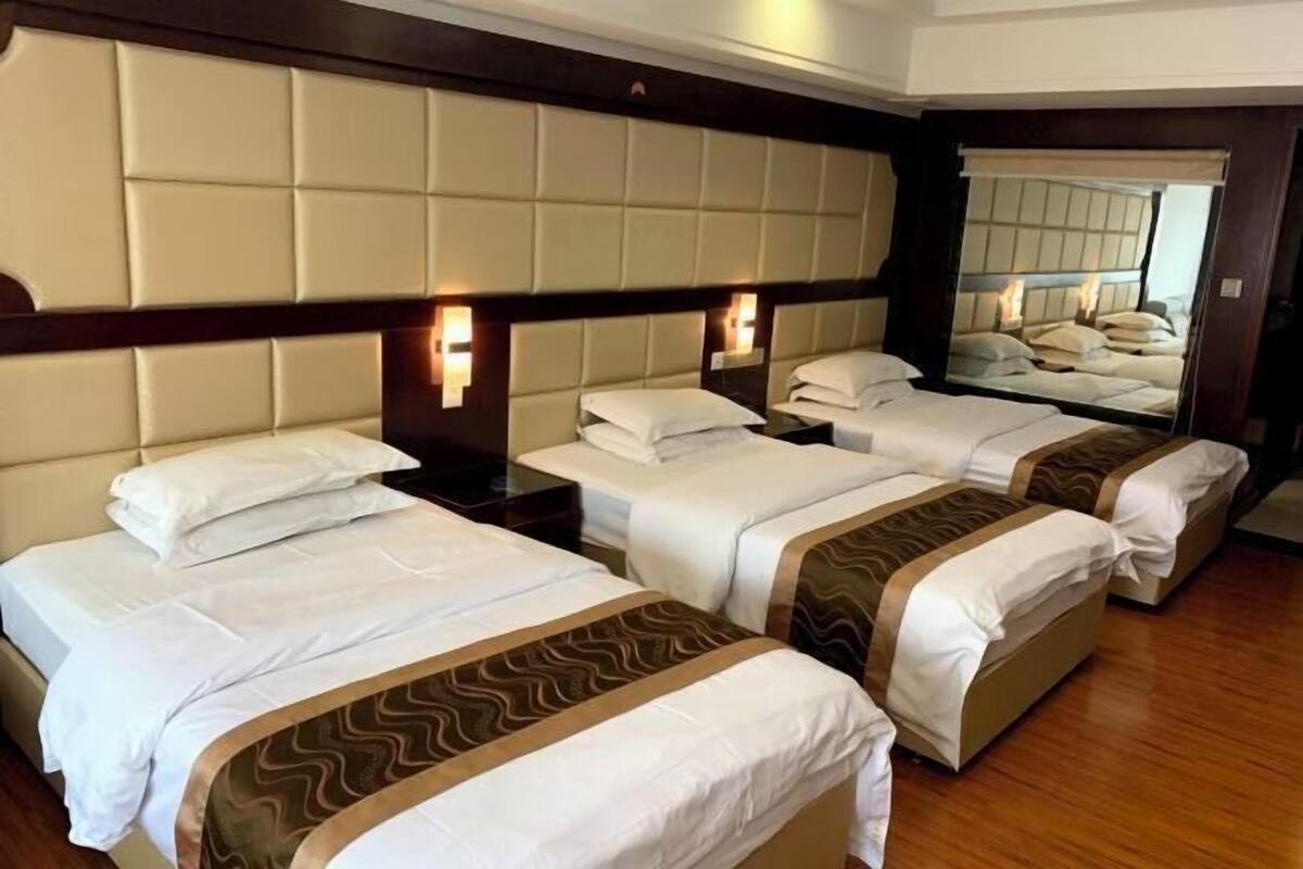 A hotel room with three beds, each with white linens and a brown decorative trim. The room features a wooden floor and a large mirror on the wall.
