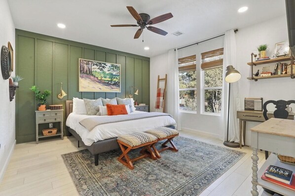 A bedroom with a bed, a desk, a chair, a ceiling fan, a window with curtains, and a bookshelf.