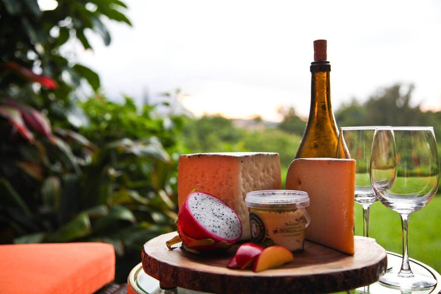 A wooden board with cheese, a bottle, a jar, and a fruit on an outdoor table with glasses.