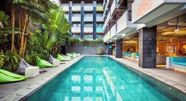 A swimming pool with lounge chairs and greenery, surrounded by a building with multiple floors and balconies.