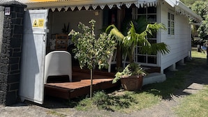 Uma pequena casa branca com telhado inclinado, varanda com toldo amarelo e placa indicando uma área de 'Perigo: Mantenha-se Afastado'.