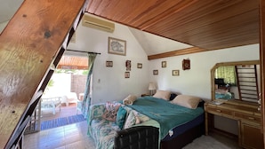 Quarto com teto inclinado, cama com colcha verde, escrivaninha de madeira e um espelho.