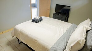 A neatly made bed with a white comforter and striped pillows, a television on a black stand, and a wooden door leading to another room. - Lake Cabin on North Long Lake (Brainerd)