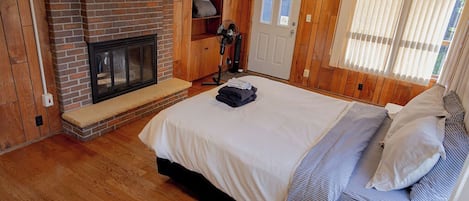 Un dormitorio con chimenea de ladrillo, una cama con manta a rayas, un banco y una ventana con persianas.