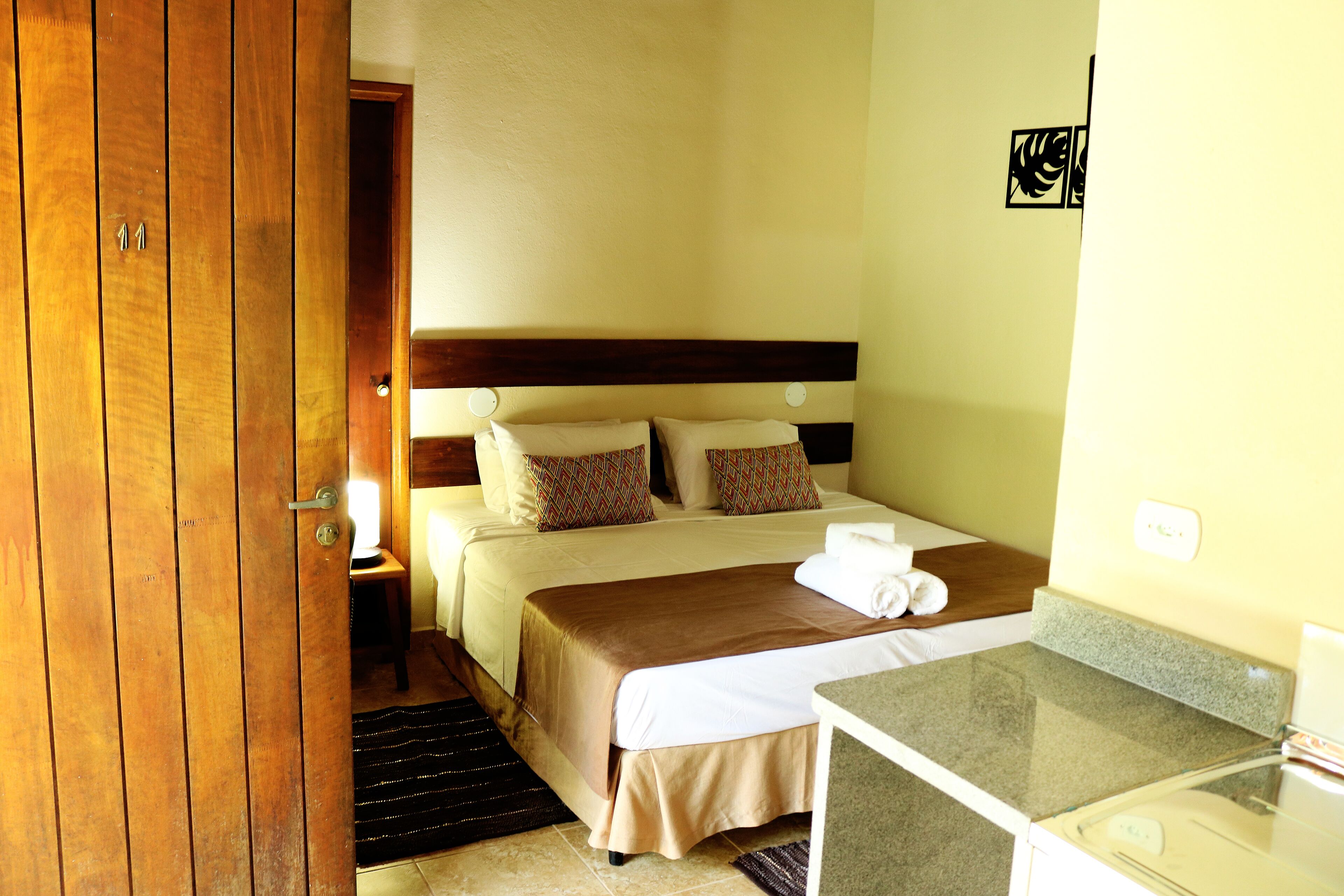 A hotel room with a wooden door, a bed with a brown headboard, a nightstand with a lamp, and a small table with a towel.