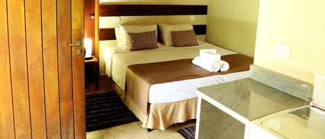 A hotel room with a wooden door, a bed with a brown headboard, a nightstand with a lamp, and a small table with a towel.