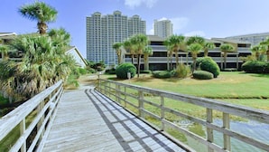 A wooden boardwalk leading to a building complex with multiple floors and a grassy area with palm trees. - 🌊 Gulf Views~Prime Spot~Pet Friendly~Steps to Beach~Vacay with a View 🌊 (Miramar Beach)