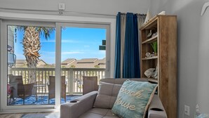A living room with a beige sofa, a blue cushion with "Beach House" text, a wooden bookshelf, and a view of a patio with outdoor furniture and a palm tree. - 🌊 Gulf Views~Prime Spot~Pet Friendly~Steps to Beach~Vacay with a View 🌊 (Miramar Beach)
