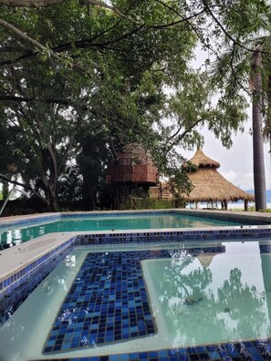 Une piscine dotée d’une terrasse carrelée, entourée d’arbres et bordée d’une structure au toit de chaume.