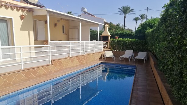A swimming pool with a tiled deck, a covered patio area, and a lush green hedge.