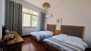 Ein Zimmer mit zwei Betten, einem hölzernen Schreibtisch mit Wasserkocher, einem Fenster mit Vorhängen und einer Pendelleuchte.